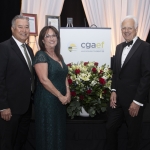 Three people in formal attire stand smiling beside a flower arrangement and a CGAEF sign at an event with white drapes and framed certificates in the background.