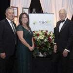 Three people in formal attire stand smiling beside a flower arrangement and a CGAEF sign at an event with white drapes and framed certificates in the background.