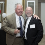 Two men in suits smile at the camera, one holding a glass of red wine with his arm around the other. They are indoors at a formal event, with balloons and a small table visible in the background.