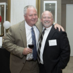 Two men in suits smile at the camera, one holding a glass of red wine with his arm around the other. They are indoors at a formal event, with balloons and a small table visible in the background.
