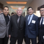 Four men in suits stand together at an indoor event, smiling at the camera. They are wearing name tags, and the background shows other people in conversation and modern decor.