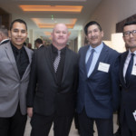 Four men in suits stand together at an indoor event, smiling at the camera. They are wearing name tags, and the background shows other people in conversation and modern decor.