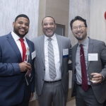 Three men in suits smile at an indoor event, each holding a drink. Behind them are white and red balloons and a table with name tags. They wear name badges and appear to be at a formal gathering or conference.