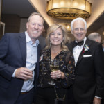 Three people dressed in formal attire smile at the camera during a social event. The woman in the center holds a glass of wine. Other guests mingle in the background under warm lighting.