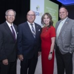 Four people in business attire pose and smile at an indoor event. A screen behind them displays WELCOME TO THE 2025 CGAE GALA OF ACHIEVEMENT DINNER with a sun logo and the acronym cgae.