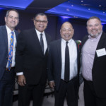 Four men in suits stand together and smile for a photo at a formal indoor event with blue lighting and decorated tables in the background.