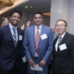 Three men in suits stand together at an indoor professional event, smiling at the camera and wearing name tags. Other attendees and event decorations are visible in the background.