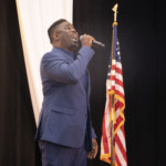 A man wearing a blue suit passionately sings into a microphone on stage, with an American flag and draped white fabric in the background.