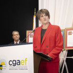 A woman in a red blazer speaks at a podium labeled cgaef California Grocers Association Educational Foundation, while a man in a suit stands nearby. Framed documents and an American flag are visible in the background.