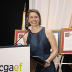A woman in a sleeveless blue dress smiles while standing at a podium with a microphone. Behind her are framed documents on easels and white drapery. The podium displays the logo “cgaef.”.