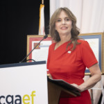 A woman in a red dress stands at a podium with cgaef written on it, smiling and holding papers. Framed documents are displayed in the background.