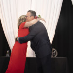 A woman in a red dress and a man in a dark suit embrace on stage in front of a black and white draped backdrop, with trophies or awards visible on a table behind them.