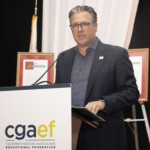 A man wearing glasses and a dark suit stands at a podium labeled cgaef California Grocers Association Educational Foundation and speaks into a microphone, with framed certificates displayed in the background.
