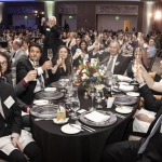 A large group of people in formal attire raise glasses for a toast around a decorated banquet table at a crowded gala or ceremony in a spacious, elegant hall.