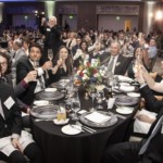 A large group of people in formal attire raise glasses for a toast around a decorated banquet table at a crowded gala or ceremony in a spacious, elegant hall.