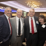 Four people dressed in business attire smile and pose together at a formal indoor event, with tables and decorative lighting visible in the background.