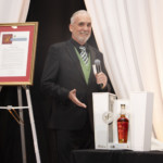 A man in a suit stands smiling at a podium with a microphone, gesturing toward a display featuring a bottle of whiskey in an open box. Framed certificates and a wine bottle are visible on a table nearby.