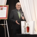 A man in a suit stands smiling at a podium with a microphone, gesturing toward a display featuring a bottle of whiskey in an open box. Framed certificates and a wine bottle are visible on a table nearby.