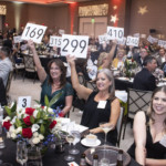 A lively auction event with attendees seated at round tables, many holding up numbered paddles to place bids. The room is decorated elegantly, and people are dressed in formal attire, smiling and engaging in the auction.