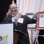 A man in a suit and glasses speaks enthusiastically at a podium labeled CGAEF, gesturing with both hands. Framed certificates and banners are visible in the background.