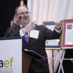 A man in a suit and glasses speaks enthusiastically at a podium labeled CGAEF, gesturing with both hands. Framed certificates and banners are visible in the background.
