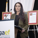 A woman with long dark hair stands at a podium labeled cgaef California Grocers Association Educational Foundation, speaking at an event with framed certificates displayed behind her.