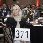 A woman sits at a banquet table holding a sign with the number 371. She is surrounded by other well-dressed people seated at round tables with floral centerpieces and dinnerware, suggesting a formal event or auction.