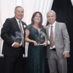 Three people stand together smiling at an awards event. Two of them, a man and a woman, are holding glass awards, while the third man stands beside them. They are dressed formally in suits and a green evening gown.