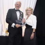 A smiling man in a tuxedo holds a glass award while standing next to a smiling woman in a white blazer and black dress, with a draped white backdrop and a wine bottle on a table behind them.
