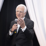 An older man with white hair, wearing a black suit and tie, holds a microphone and gestures while speaking. He stands in front of white drapes and a dark background, appearing to address an audience.