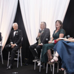 Five people in formal attire sit on a stage with white drapery and black accents. Two women and three men are talking and smiling; one man holds a microphone while the others listen and laugh.