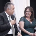 A man in a suit and tie speaks into a microphone while sitting next to a smiling woman in a green lace dress, who is also holding a microphone, at an indoor event.