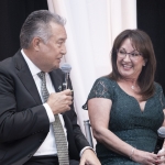 A man in a suit and tie speaks into a microphone while sitting next to a smiling woman in a green lace dress, who is also holding a microphone, at an indoor event.