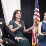 Three people sit on stage in front of an American flag. The woman in the middle speaks into a microphone while the man on the left and the woman on the right listen. All are dressed formally.