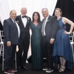 Five adults stand together smiling, dressed in formal evening attire, with a white draped backdrop and chairs behind them. The group includes three men in suits or tuxedos and two women in floor-length dresses.
