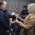 Two people smile and exchange a navy blue tote bag at an indoor event, while three others stand in the background talking and holding bags. The atmosphere appears friendly and casual.