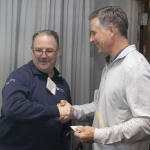 Two men are indoors, smiling and shaking hands. One wears a navy blue sweater with a name badge, while the other wears a light gray shirt and holds a notepad. Gray curtains and an open door are visible in the background.