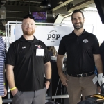 Four men stand and smile together indoors at an event booth for “double pop soda.” They wear event badges and casual business attire; golf clubs and sports screens are visible in the background.