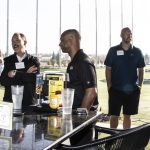 Five men stand and talk around a bar-height table at an outdoor driving range, with golf nets and a green field visible in the background. They appear to be socializing at a professional or networking event.