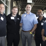 Five men stand together smiling for a group photo in an indoor-outdoor venue, wearing casual or business-casual clothing and name tags, with golf bays and screens visible in the background.