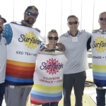 Five people stand together at a Topgolf venue, three of them wearing colorful Surfside canned drink costumes. They are smiling, and various Surfside drink products are displayed in front of them.