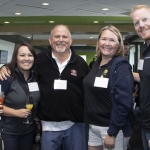 Five adults stand closely together indoors at a social event, smiling at the camera. Two women hold drinks, all wear name badges, and the group stands in a modern, well-lit room with others mingling in the background.