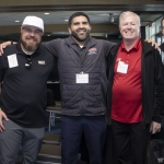 Five men stand together indoors, smiling at the camera, wearing conference name tags. Some have their arms around each other in a friendly gesture. The background shows chairs and large windows with daylight coming in.
