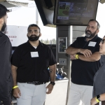 Four people stand and talk in a semi-circle at an indoor-outdoor venue with golf equipment visible. Two men wear black shirts, one man wears a jacket, and a woman wears sunglasses. Monitors and grassy scenery are in the background.
