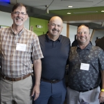 Five people stand close together indoors, smiling at the camera. They wear name tags and casual attire, suggesting a professional or social event. The background shows other attendees and modern interior features.