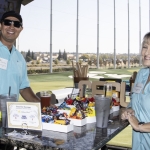 Two people wearing light blue shirts and name tags stand at a counter with snacks and drinks at an outdoor golf entertainment venue, with golf bays and nets visible in the background. Blue and red balloons are nearby.