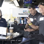 Three people stand at a high table in a golf driving range. Two men and one woman are wearing dark polo shirts with name tags. One man flashes a peace sign while the others smile or check a phone. Golf bays and screens are visible.