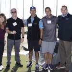 Five people stand together and smile at an outdoor driving range, with golf balls, clubs, and targets visible in the background under a sunny sky. They all wear name tags and casual or business casual clothing.