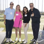 Four people stand smiling at a golf driving range, with one woman holding a golf club. They wear casual golf attire and name tags, and a man on the right gives a thumbs up. Sunlight brightens the outdoor setting.