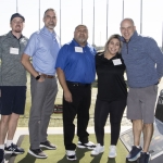 Five people stand smiling at a golf driving range, wearing name tags and casual clothes. The group poses together in front of golf bays and nets, with bright outdoor lighting and a Topgolf sign visible.